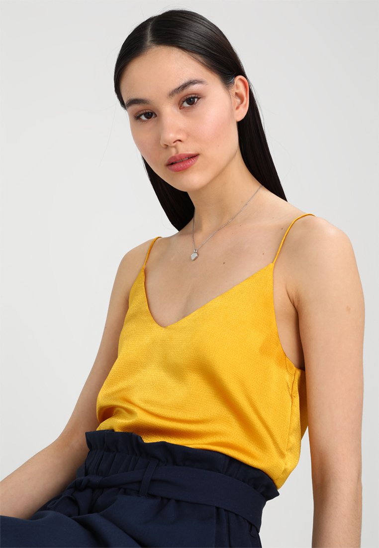 Gelbe Satin-Camisole mit dünnen Trägern und V-Ausschnitt, kombiniert mit hoch taillierten marineblauen Hosen. Glatte Textur, minimalistisches Design.