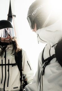 Two skiers in white and light green ski jackets with black accents, wearing black helmets and mirrored goggles, against a bright background.
