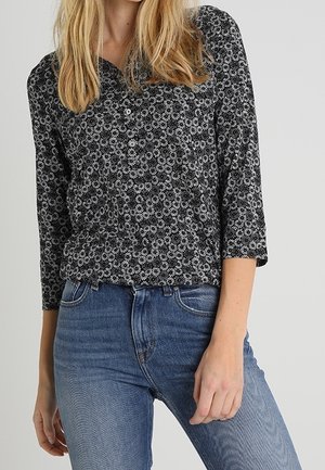 Kvinde iført sort skjorte med knapper og hvidt blomsterprint samt blå jeans med højtaljet talje, stående mod en ensfarvet baggrund.