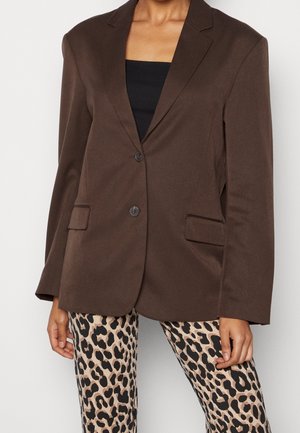 Personne portant un blazer marron à deux boutons, un haut noir et un pantalon beige à motifs léopard, debout devant un fond clair uni.