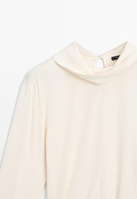 Haut à manches longues couleur crème avec un col rabattu et une fermeture à bouton au dos. Tissu lisse avec un design minimaliste et sans motifs.