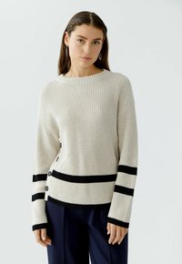 Cremfarbener Rippstrickpullover mit schwarzen Streifen am Saum und an den Ärmeln. Merkmale sind ein runder Ausschnitt, lange Ärmel und schwarze Knöpfe an der Seite.