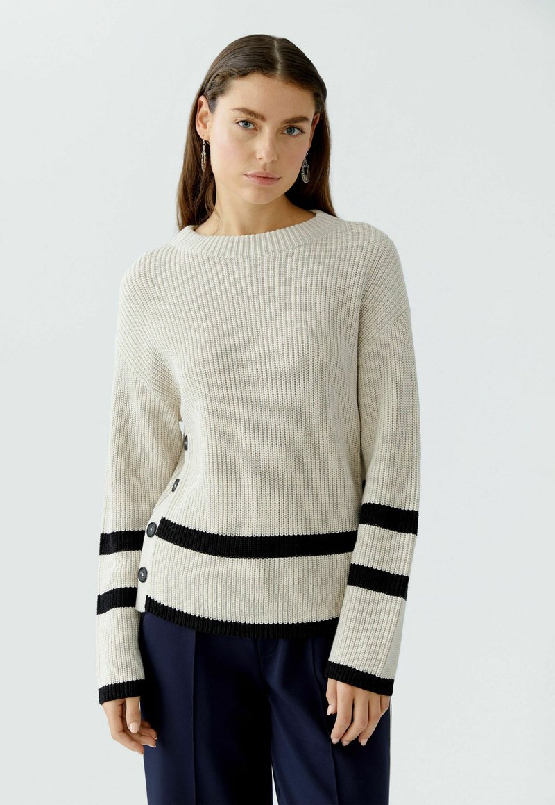 Cremfarbener Rippstrickpullover mit schwarzen Streifen am Saum und an den Ärmeln. Merkmale sind ein runder Ausschnitt, lange Ärmel und schwarze Knöpfe an der Seite.