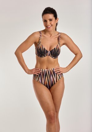 Lachende vrouw met donker haar in een gestreepte bikini met hoge taille, staand met handen in haar zij tegen een effen lichte achtergrond.