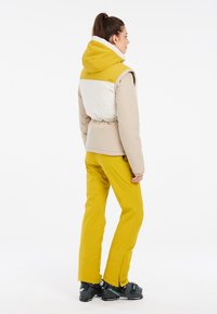 Bežinė ir balta pūkinė striukė su geltonu kapišonu, susiaurinta juosmenyje. Derinama su geltonomis vandeniui atspariomis kelnėmis ir juodais slidinėjimo batais.