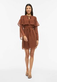 Robe marron à pois avec des volants à étages, manches longues et ceinture à la taille. Tissu transparent qui ajoute de la texture. Sandales noires complètent le look.