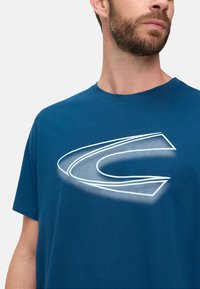 Blue cotton t-shirt featuring a large white graphic design with gradient accents. Short sleeves and a crew neckline.