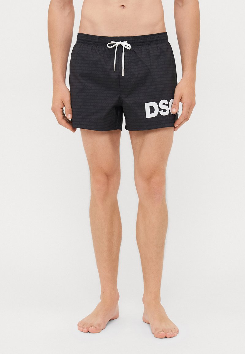 Mann trägt schwarze Badeshorts mit weißem Kordelzug und großen weißen Buchstaben "DSQ" auf dem rechten Oberschenkel, steht barfuß vor einem weißen Hintergrund.