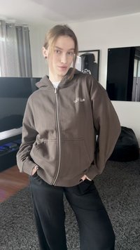 Sweat à capuche marron avec fermeture éclair, doté d'un logo "Cozy Club" sur la poitrine gauche. Porté avec un pantalon noir, sur un tapis gris texturé.