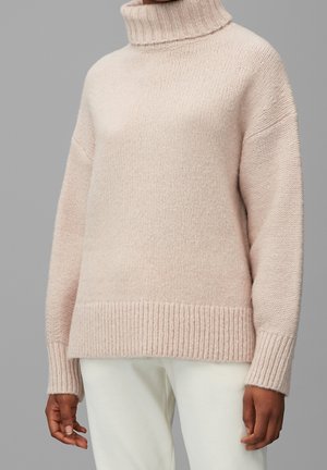 Personne portant un pull en maille à col roulé beige clair associé à un pantalon blanc cassé, debout devant un fond gris.
