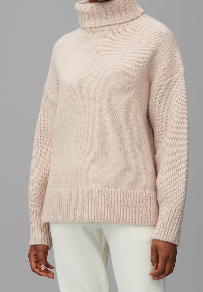 Personne portant un pull en maille à col roulé beige clair associé à un pantalon blanc cassé, debout devant un fond gris.