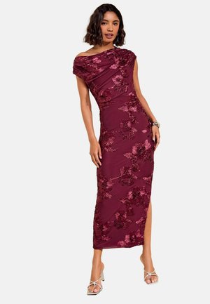Kvinde iført en vinrød off-shoulder kjole med blomsterbroderi og sidelommer, kombineret med sølvfarvede højhælede sandaler og et armbånd.