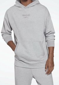 Homme portant un sweat à capuche Reebok gris clair et un pantalon de survêtement assorti, une main dans la poche du sweat et l'autre reposant le long du corps.