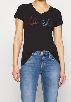 Kobieta w czarnym T-shircie z dekoltem w serek z czerwonym i niebieskim tekstem z cyrkonii oraz w niebieskich jeansach z zapięciem na guzik i zamek błyskawiczny