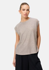 Kobieta z zaczesanymi do tyłu włosami, ubrana w błyszczącą beżową bluzkę bez rękawów i czarne spodnie, stoi z jedną ręką w kieszeni na jednolitym tle.