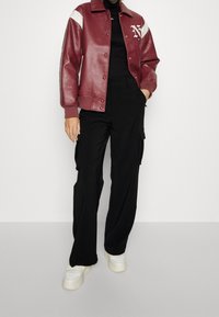 Bordeauxfarbene Kunstlederjacke mit weißen Akzenten und Logo, kombiniert mit schwarzen Cargo-Hosen und weißen Sneakern. Modernes, sportliches Design.