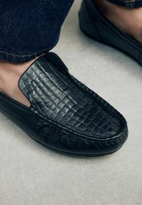 Chaussures noires en cuir à enfiler avec une texture crocodile, bout rond et détails cousus le long de la semelle, associées à un jean en denim foncé.