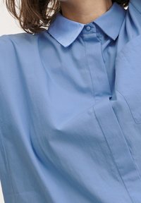 Camicia in cotone azzurro chiaro con colletto classico, texture liscia e chiusura a bottoni. Presenta una tasca sul petto e una vestibilità comoda.