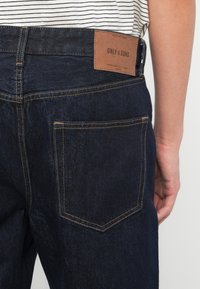 Κοντινό πλάνο ατόμου που φοράει σκούρο μπλε τζιν με δερμάτινο ταμπελάκι της μάρκας "Only & Sons" σε ταμπά χρώμα πάνω από την πίσω τσέπη.