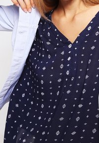 Mørkeblå mønstret bluse med hvide geometriske designs, v-hals, letvægtsstof og en afslappet pasform. Knapper og korte ærmer synlige.