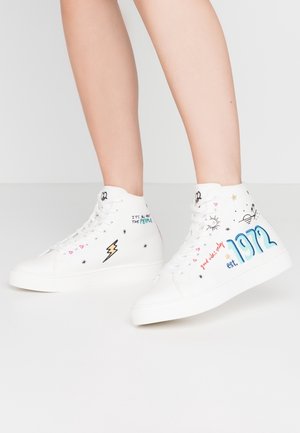 Jambes portant des baskets montantes blanches ornées de gribouillis colorés, comprenant "1972", des étoiles, un éclair et des motifs de soleil sur un fond uni.