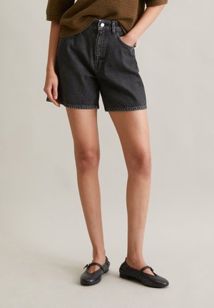 Zwarte denim shorts met een hoge taille, vijf zakken en een rauwe zoom. Gemaakt met zwarte platte schoenen. Achtergrond is eenvoudig en neutraal.
