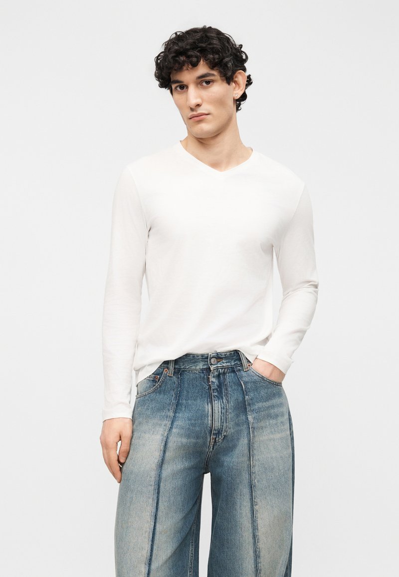 Jonge man met krullend haar, gekleed in een wit langärmelig shirt met V-hals en losse, tweekleurige blauwe spijkerbroek, staand tegen een witte achtergrond.