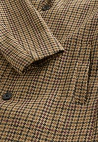 Pruunid ja mustad hundhammasmustriga kangas, millel on peened bordoopunased aktsendid, nööpidega suletav ja volditud krae detailiga.