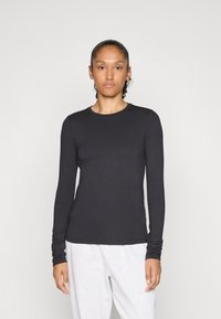 Frau mit zurückgegebem Haar trägt ein eng anliegendes schwarzes Langarmshirt und hellgraue Hosen und steht vor einem schlichten, hellen Hintergrund.