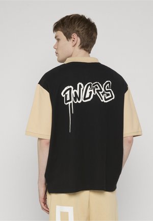 Junger Mann im Profil, der ein kurzärmeliges Hemd in Schwarz und Beige trägt, mit weißem Graffiti-Text "DNGRS" auf der Rückseite.