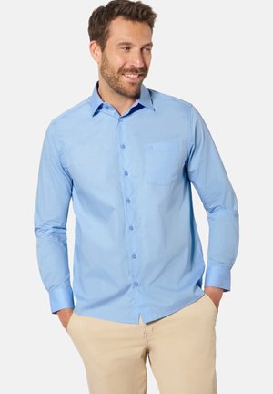 Man with short brown hair and beard wearing a light blue button-up shirt and beige pants with hands in pockets, looking to the side smiling.