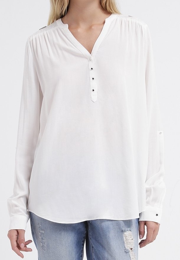 Chemisier blanc à manches longues avec un décolleté en V, patte de boutonnage et épaules froncées. Tissu doux et coupe décontractée, associé à un jean bleu délavé.