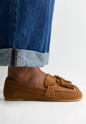 Mocasín marrón de ante con borlas, usado con vaqueros azules remangados y pies descalzos sobre fondo blanco.