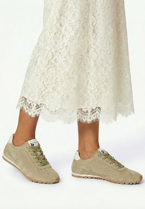 Benen die beige suède sneakers met veters en witte accenten dragen, gecombineerd met een witte kanten midi-rok tegen een witte achtergrond.