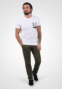 Hvit t-skjorte med svart palmetre-grafikk på brystlommen, kombinert med olivengrønne bukser og svarte sko. En enkel, uformell antrekk.