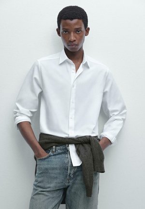 Weißes Hemd mit umgeschlagenen Ärmeln, kombiniert mit hellblauen Jeans. Ein dunkles Strickmaterial dient als Taillengürtel.