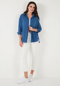 Camisa azul de botones con un acabado texturizado, mangas remangadas, combinada con pantalones cortos blancos y zapatillas blancas. Atuendo simple y casual.
