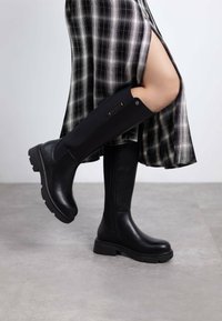 Botas negras hasta la rodilla con un panel texturizado negro, suela gruesa y detalles de hardware en los lados. Combinadas con una falda de cuadros.