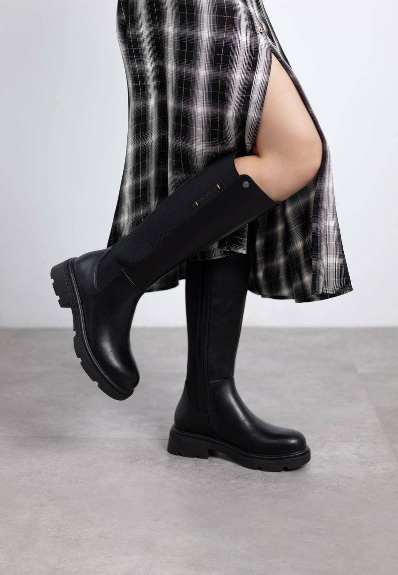 Botas negras hasta la rodilla con un panel texturizado negro, suela gruesa y detalles de hardware en los lados. Combinadas con una falda de cuadros.