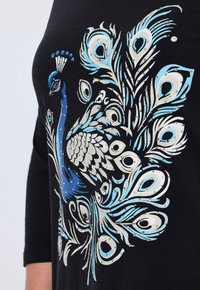 Schwarzes Shirt mit einem Pavo-Design, das blaue und silberne Akzente, komplizierte Feder-Muster und Strassverzierungen aufweist.