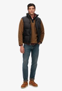 Svart pufferväst över en brun fleece-tröja, kombinerad med blå jeans och beige stövlar. Outfiten har en rutigt skjorta under.