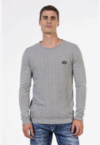 Grauer Strickpullover mit einem strukturierten Zickzackmuster, rundem Ausschnitt und gerippten Bündchen. Kleines Logopatch sichtbar auf der Brust.
