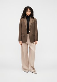 Blazer marron sur mesure avec deux poches avant, fermeture à un bouton ; associé à un pantalon beige à jambes larges et un col roulé noir.