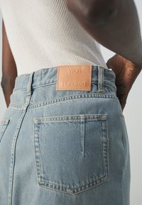 Gros plan sur une personne portant un jean en denim bleu clair avec un patch en cuir beige marqué "Won Hundred" et un haut côtelé beige.