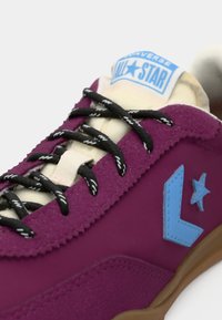 Purple suede sneaker features black and white striped laces, a light blue logo, and a cream-colored tongue. Textured rubber sole visible.