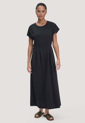 Frau trägt ein schlichtes schwarzes Maxikleid mit kurzen Ärmeln und dunkelgrüne Sandalen, steht vor einem einfarbigen hellen Hintergrund.