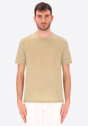 Uomo in piedi che guarda avanti, indossa una maglietta beige a maniche corte e pantaloni bianchi su uno sfondo uniforme.