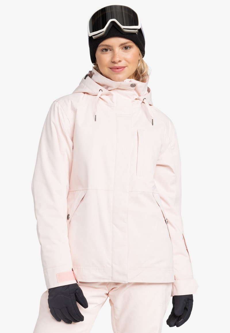 Lichtroze skijack met een capuchon, voorzien van ritszakken, verstelbare manchetten en een gladde textuur. Zwarte handschoenen en een skibril complementeren de outfit.