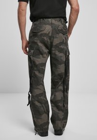 Pantalones cargo en un patrón de camuflaje oscuro, fabricados con tela resistente. Incluyen múltiples bolsillos laterales, tirantes ajustables y un corte holgado.