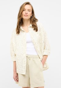 Lightweight, long-sleeve button-up shirt in pale cream with leaf patterns, paired with beige shorts featuring an elastic waistband and pockets.